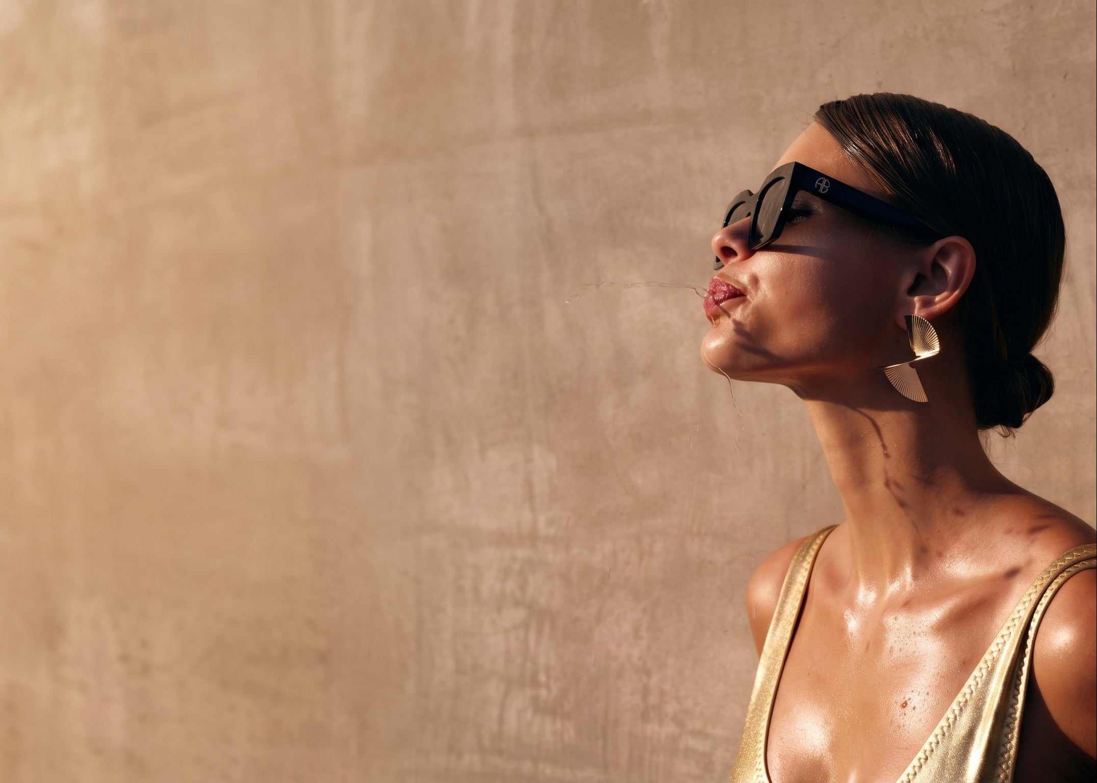 Woman wearing sunglasses and a gold top against a beige wall