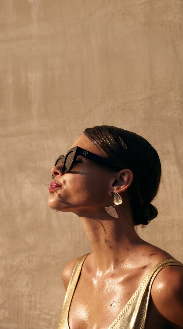 Woman wearing sunglasses and a gold top against a beige wall
